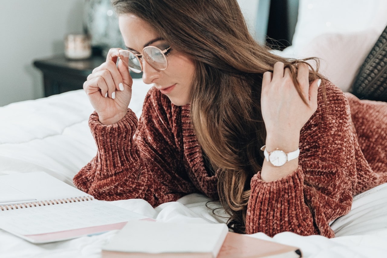 Woman wearing glasses and a fuzzy sweater laying on her bed looking at notebooks while wearing a white minimalist watch and white nail polish