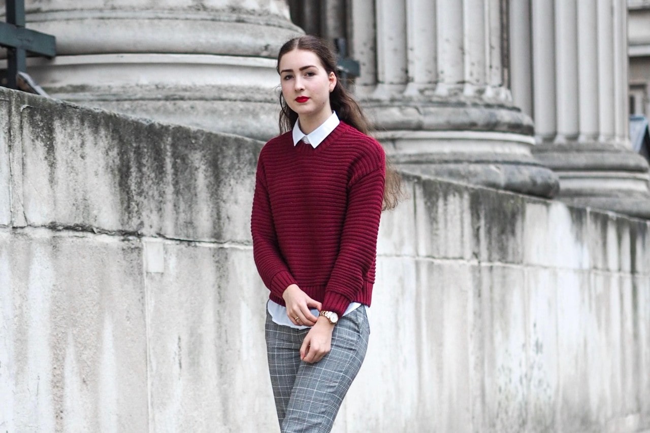 Woman walking in front of a building in a maroon sweater and gray plaid pants adjusting her sterling silver watch