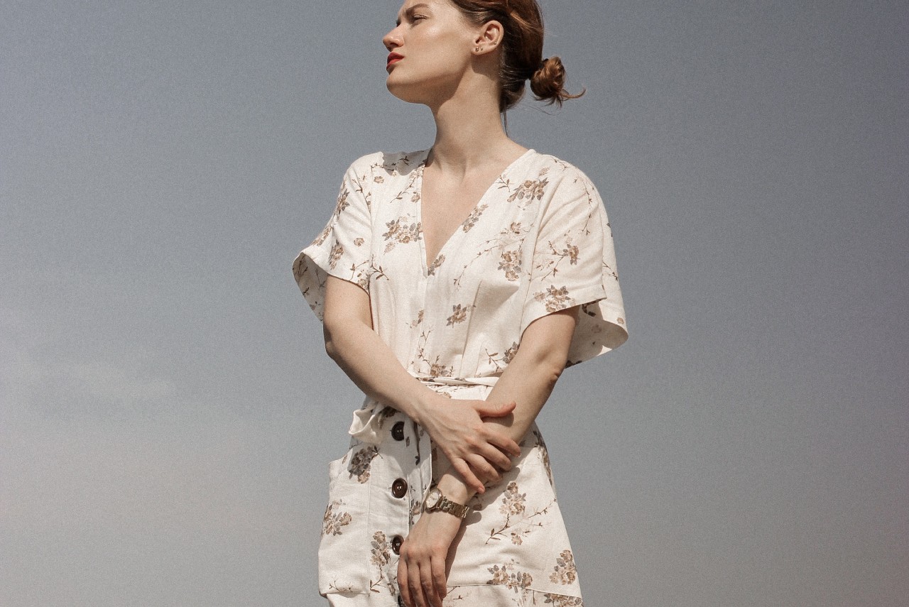 Standing in the sun against a clear blue sky is a woman in a white sundress with a floral pattern and gold watch