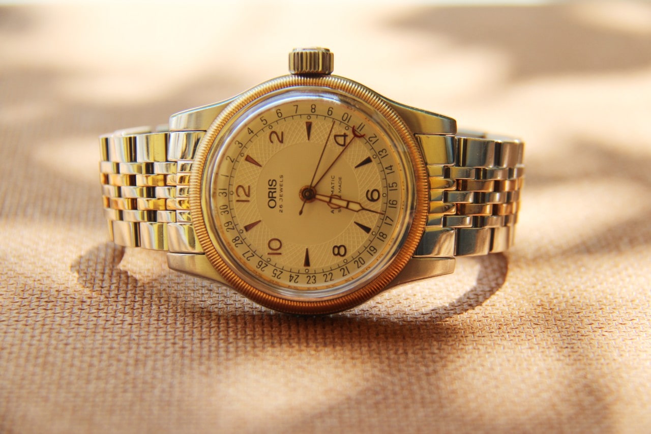 An 18k yellow gold watch from Oris sits on a gold-colored fabric table cloth during an outdoor picnic
