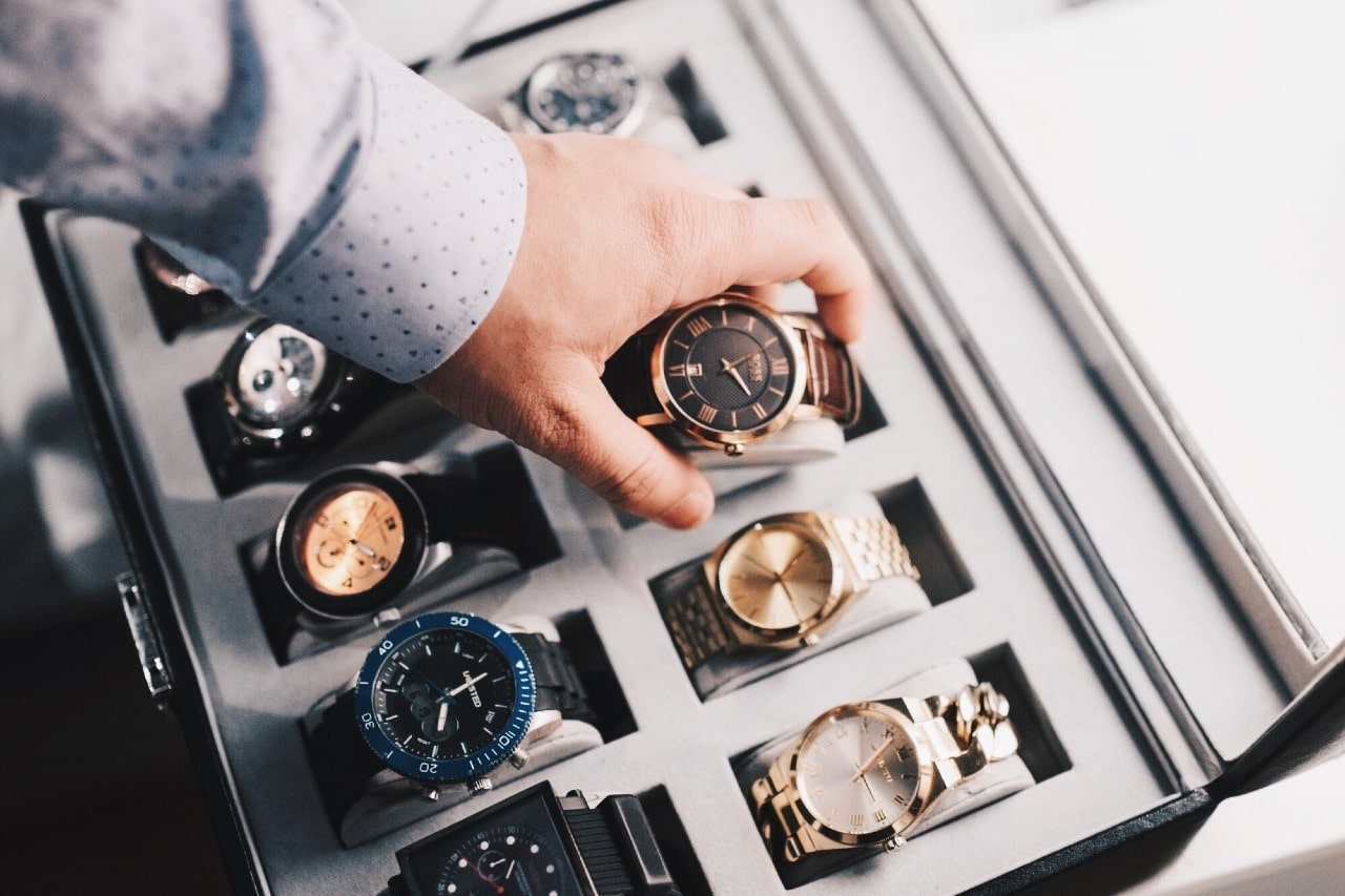 A handsomely dress arm picks up and inspects a timepiece out of a case full of timepieces