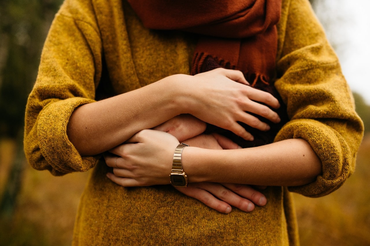 A woman in a yellow sweater and gold watch, with her partner’s arm wrapped around her