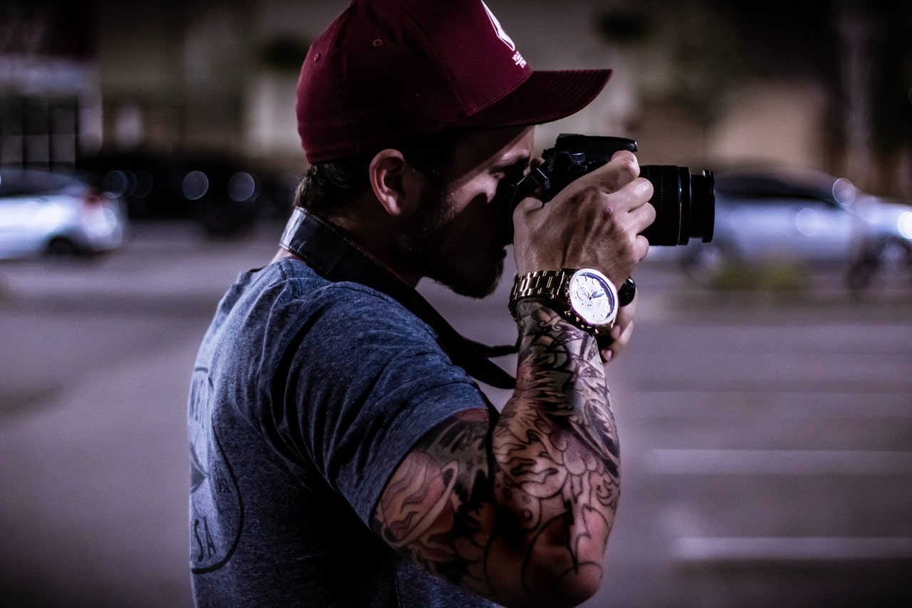 A tattooed photographer in a parking lot shows off his watch.