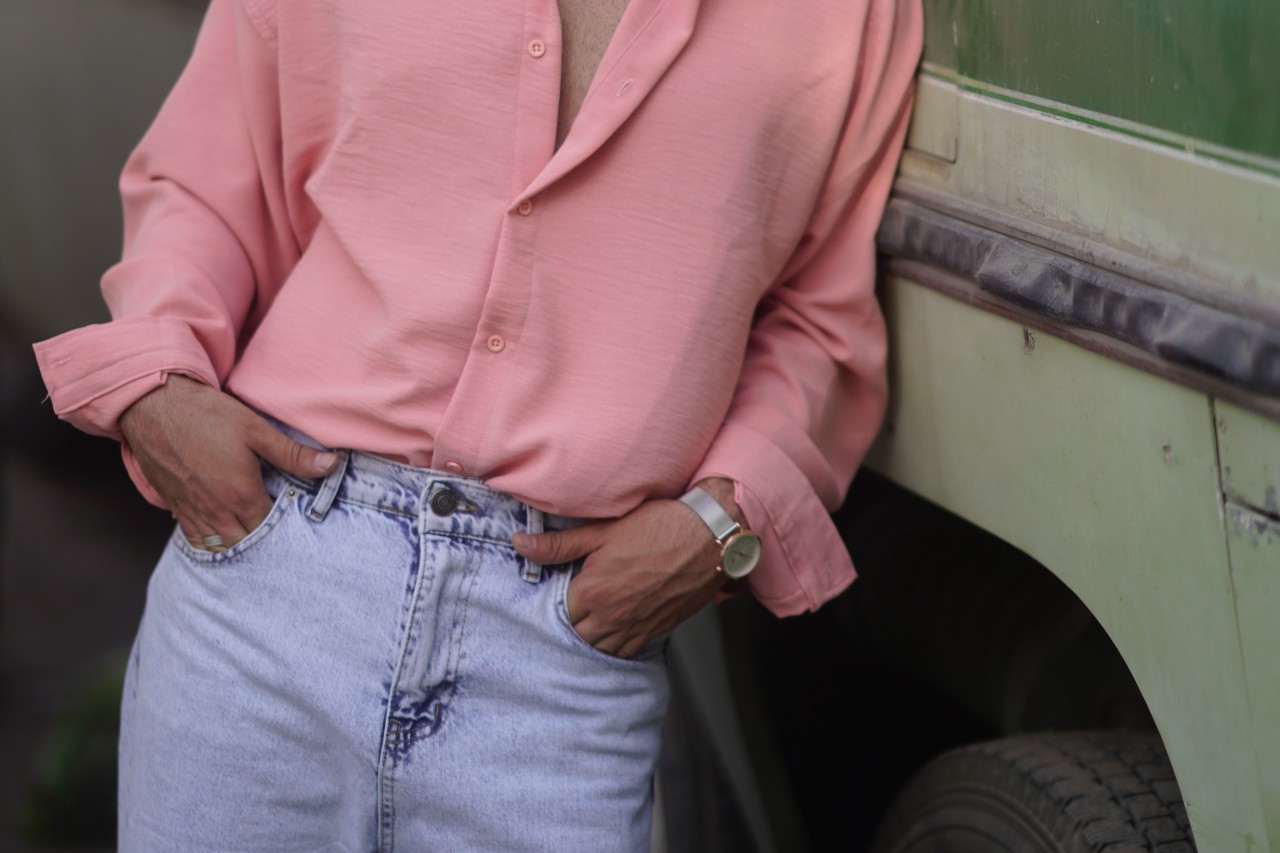 a man leaning against a truck, wearing a pink top and jeans and a silver watch