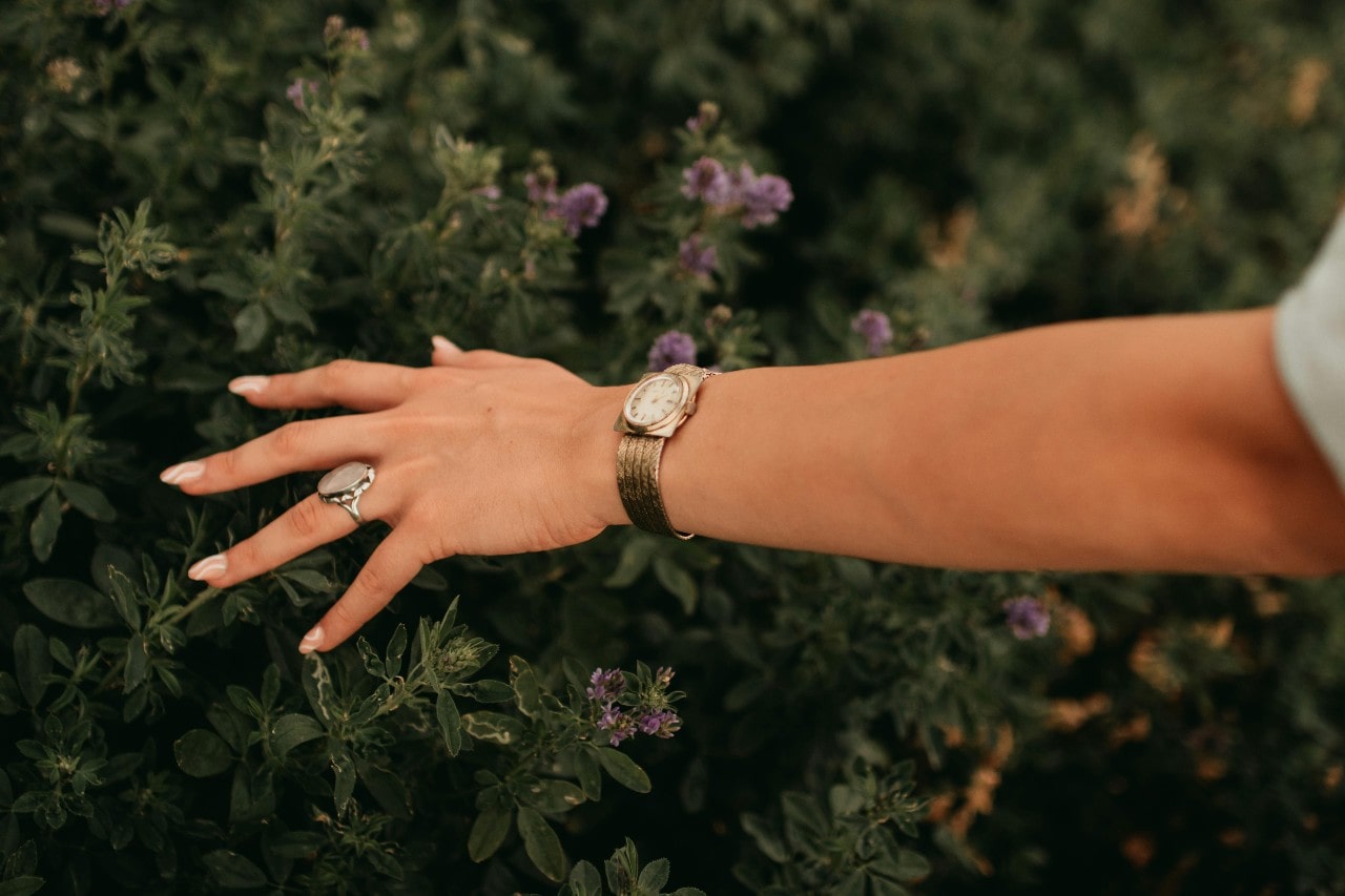 a woman’s extended hand, touching flowers and wearing a yellow gold watch