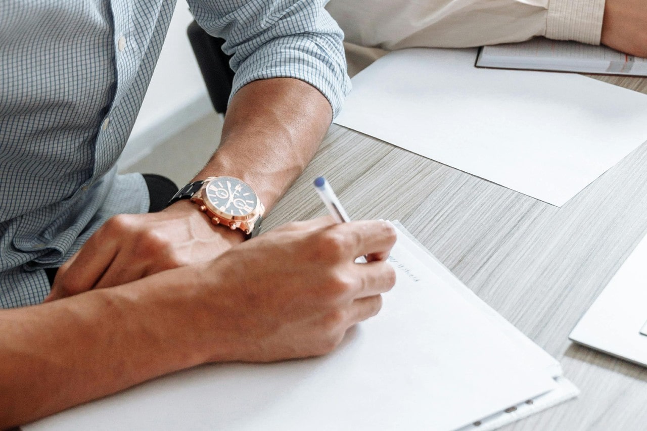 A close-up image of a man writing at a desk and wearing a luxury watch.