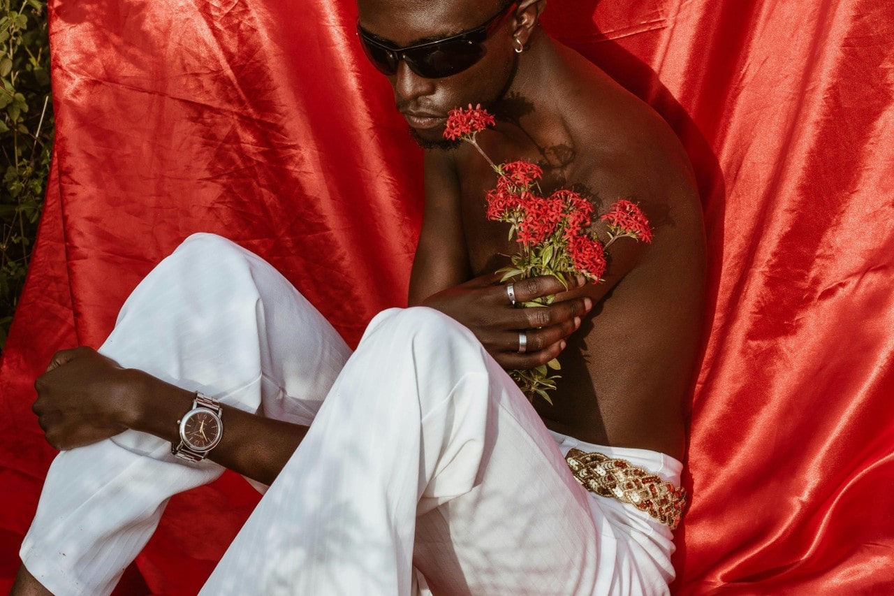 A man posing in front of a red satin sheet, holding red flowers and wearing a luxury watch.