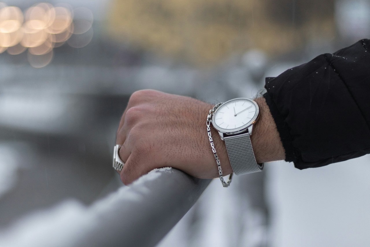 a man’s hand resting on a rail, adorned with a silver watch and chain bracelet and ring