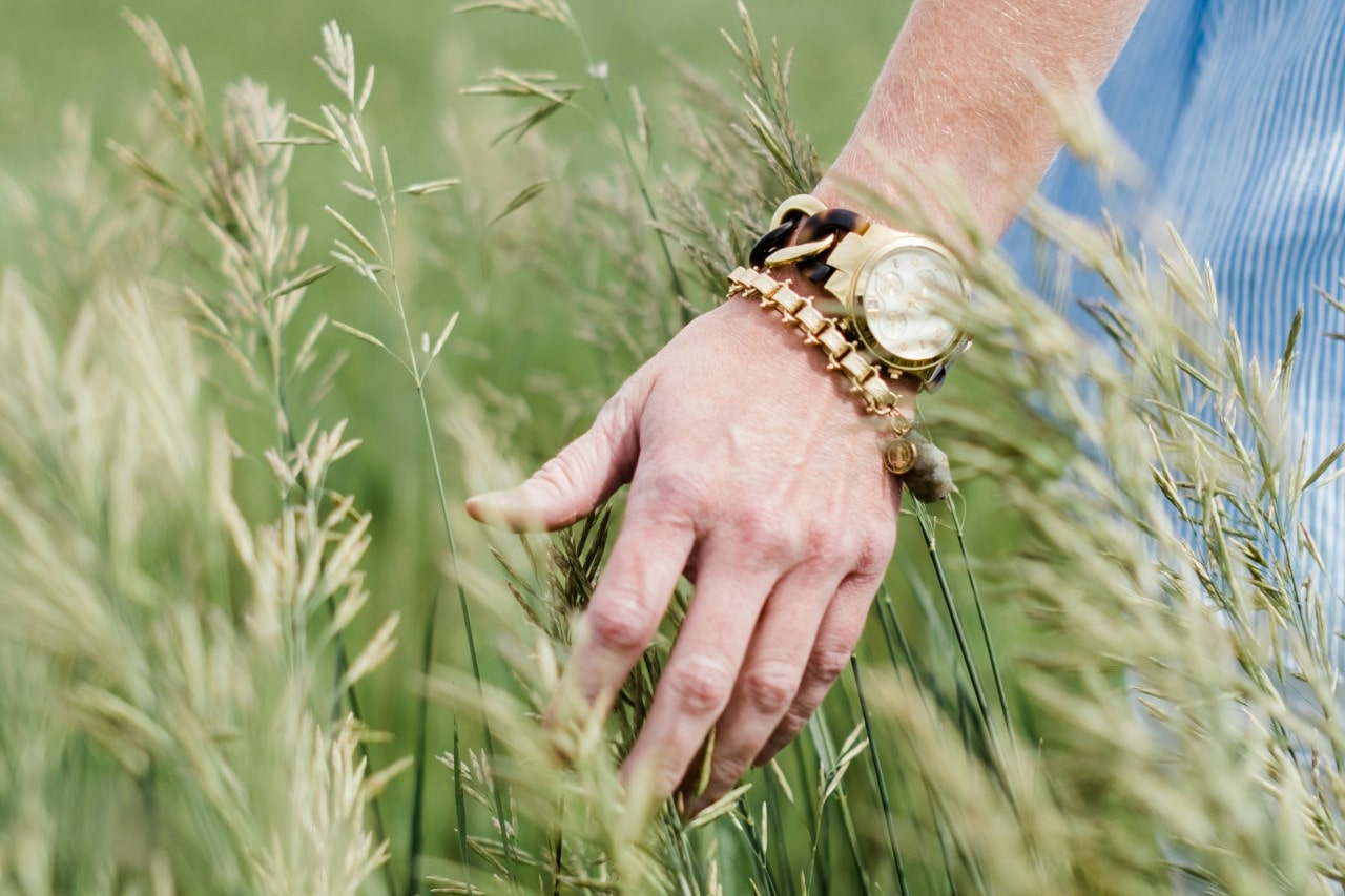 a woman’s arm touching tall grass and wearing a watch and large gold chain bracelet