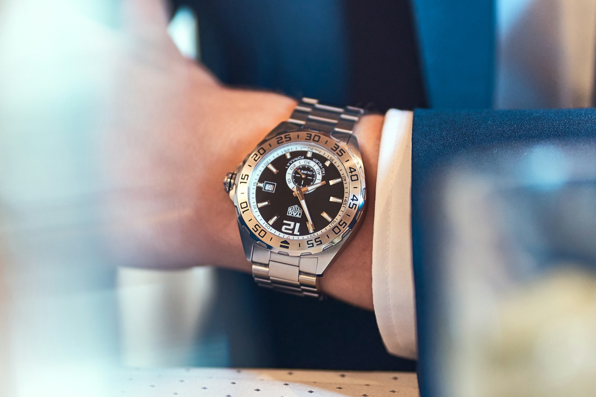 A close-up of a well-dressed man in a blue suit, wearing a sporty TAG Heuer watch.