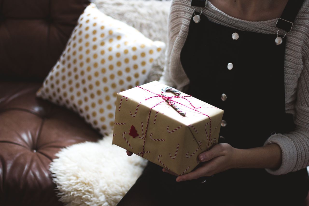 A close-up of a young woman holding a wrapped gift with a candy cane tied on top, waiting to present it to her partner.