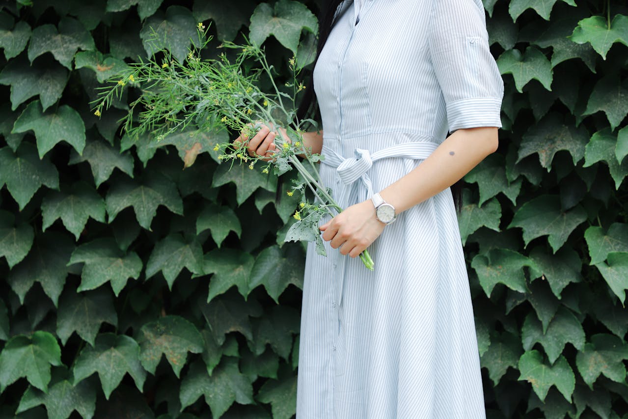 A photo of a woman wearing a spring dress and a luxury watch standing in front of a wall of ivy, holding a large sprig of greenery.