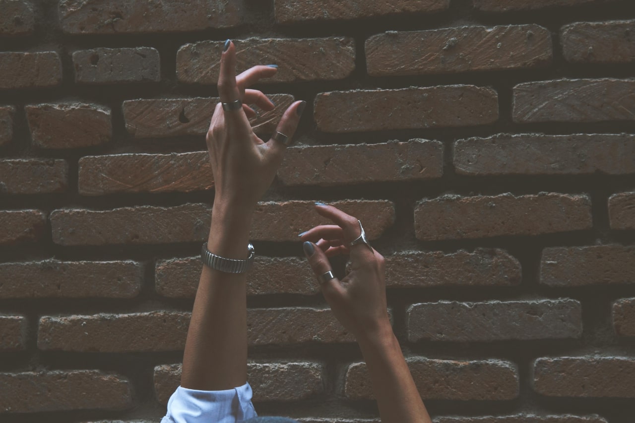 A close-up of a woman’s hands adorned with a fashionable watch and jewelry, reaching toward the sky before a brick wall.