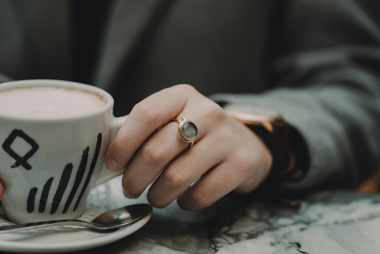 A woman wearing a watch and a ring sips coffee from a unique mug.