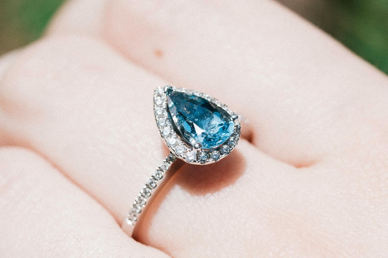 close up image of a hand adorned with a white gold ring featuring a blue pear shaped aquamarine with a halo of diamonds around the center stone and around the band