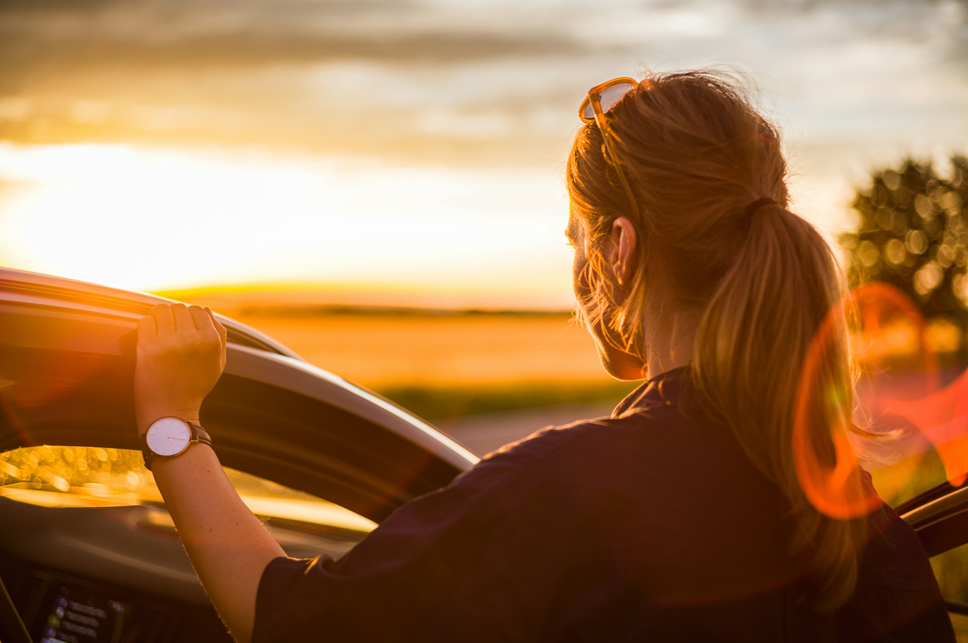 a lady wearing a watch resting against her car looking at a sunset