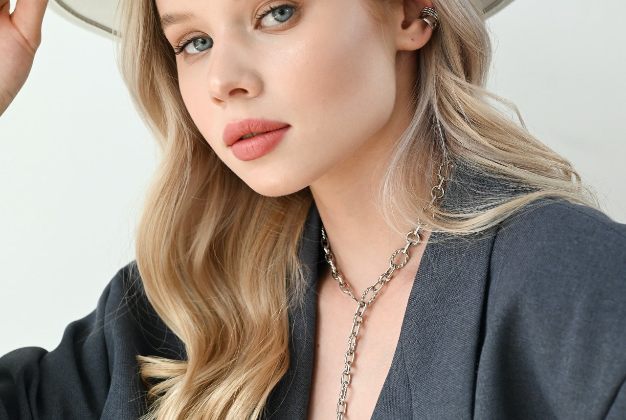 A close-up photo of a pretty young woman wearing a distinctive figaro chain necklace, peering at the camera from under her hat brim.