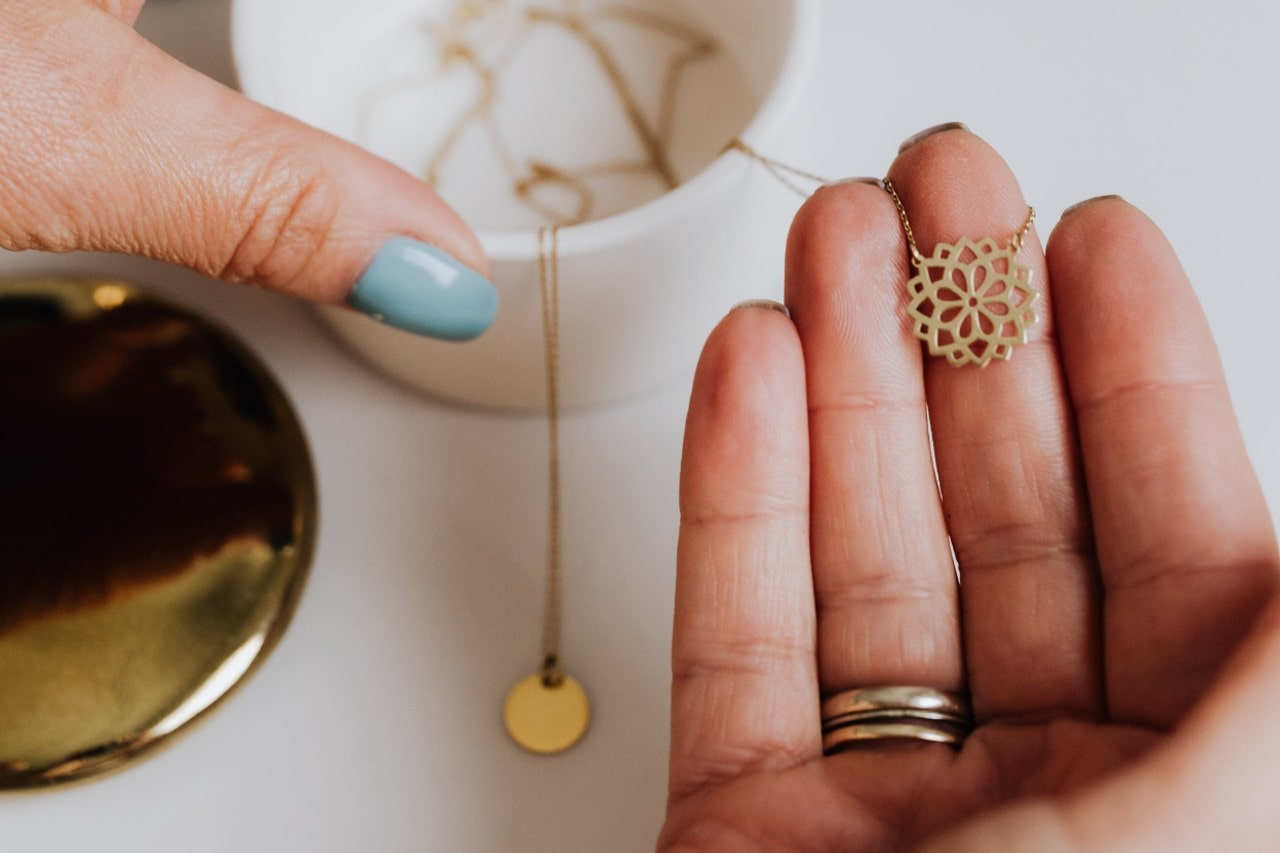 A close-up of a woman’s hand selecting a delicate gold pendant necklace from a small jewelry box.